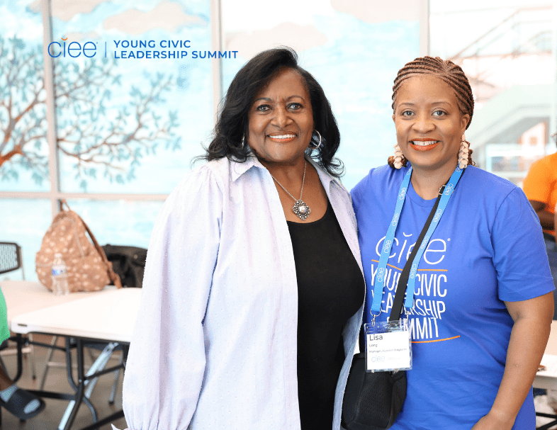 Two middle-aged women stand in a brightly lit room wearing blue shirts that say "Young Civic Leadership Summit"