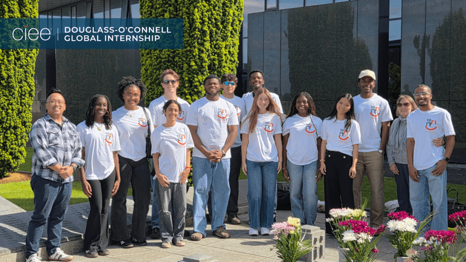 The cohort gathers outdoors at a memorial site surrounded by flowers and modern stone structures. Most of the students are wearing matching white program T-shirts, creating a unified look. Two adults stand at either end of the group, likely program leaders or hosts. The setting suggests a cultural or historical visit, emphasizing the educational and intercultural components of the internship program alongside the students’ professional development experiences.